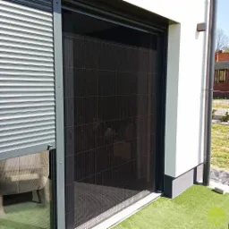 Zewnętrzna, plisowana moskitiera w kolorze czarnym zamontowana na oknie tarasowym, obok zwiniętej rolety zewnętrznej w kolorze srebrnym, na tle białej ściany budynku.