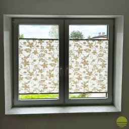 MARKIZA.PL SPÓŁKA Z OGRANICZONĄ ODPOWIEDZIALNOŚCIĄ - Dwuskrzydłowe okno z białymi ramami, osłonięte plisowanymi roletami z motywem kwiatowym, widok na zielony trawnik i drzewa.