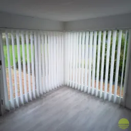 Wnętrze jasnego pomieszczenia z dużymi oknami zasłoniętymi białymi, pionowymi żaluzjami materiałowymi; widok na zielony ogród za oknem.