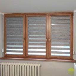 MARKIZA.PL SPÓŁKA Z OGRANICZONĄ ODPOWIEDZIALNOŚCIĄ - Trzy okna z drewnianymi ramami wyposażone w szare rolety z poziomymi pasami, widoczny grzejnik pod oknem.