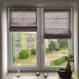 MARKIZA.PL SPÓŁKA Z OGRANICZONĄ ODPOWIEDZIALNOŚCIĄ - Dwa okna PCV z szarymi roletami rzymskimi, widok na zewnątrz, dekoracje na parapecie: klepsydra, świeczniki i bonsai.