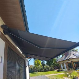 MARKIZA.PL SPÓŁKA Z OGRANICZONĄ ODPOWIEDZIALNOŚCIĄ - Zewnętrzna, rozkładana markiza w kolorze grafitowym zamontowana nad oknem z żaluzjami, widok z boku na tle błękitnego nieba i zadbanego ogrodu z domem.