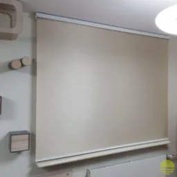 Beżowa roleta zwinięta do połowy okna, widoczna aluminiowa kaseta i dolna listwa, na ścianie obok drewniane dekoracje.