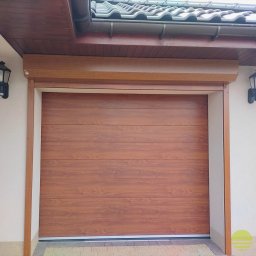 MARKIZA.PL SPÓŁKA Z OGRANICZONĄ ODPOWIEDZIALNOŚCIĄ - Zewnętrzna brama garażowa segmentowa w kolorze drewna, z widoczną strukturą poziomych paneli, zamontowana w jasnej elewacji budynku z ciemnobrązowym dachem.