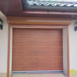 Zewnętrzna brama garażowa segmentowa w kolorze drewna, z widoczną strukturą poziomych paneli, zamontowana w jasnej elewacji budynku z ciemnobrązowym dachem.