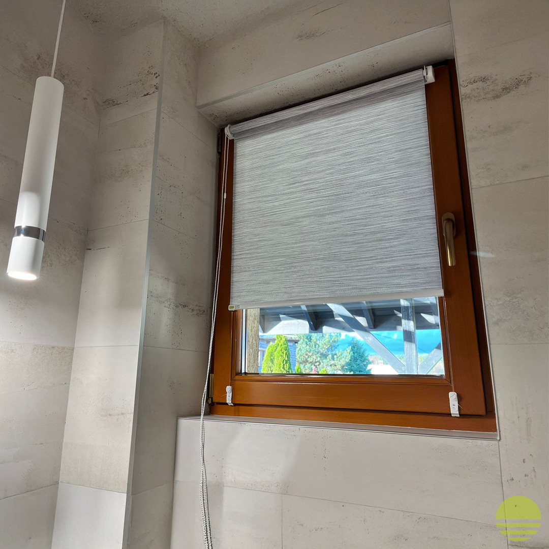 Jasnoszara roleta zwijana z delikatnym wzorem na brązowym oknie PCV, widok na zewnątrz z zielenią i błękitnym niebem, nowoczesna lampa wisząca w tle, marmurowe płytki na ścianie.