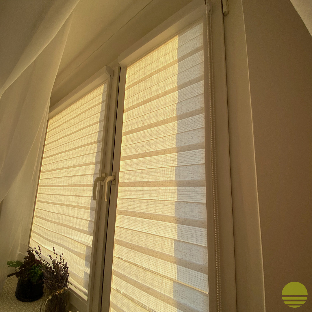 Beżowe, poziome pasy rolety okiennej dzień noc, częściowo zasłoniętej, z widocznym mechanizmem łańcuszkowym, w oknie PCV, z dekoracją roślinną na parapecie, oświetlone ciepłym światłem.