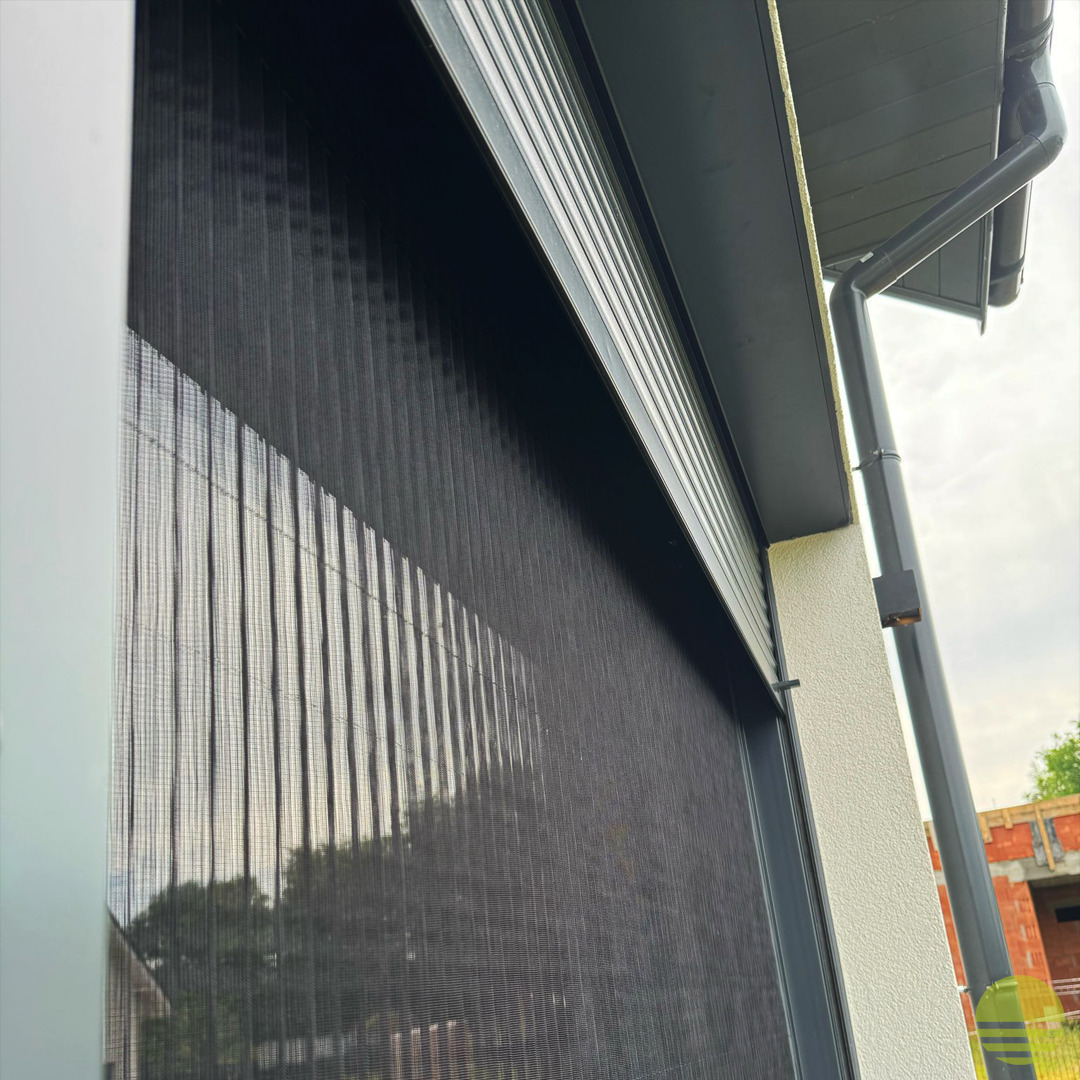 Czarna, zwijana przesłona okienna z widoczną strukturą siatki, zamontowana na elewacji budynku z rynną spustową. W tle zarys drzew i budowa.