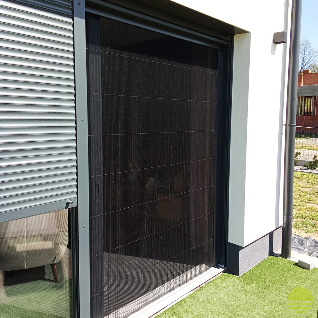 Zewnętrzna, plisowana moskitiera w kolorze czarnym zamontowana na oknie tarasowym, obok zwiniętej rolety zewnętrznej w kolorze srebrnym, na tle białej ściany budynku.