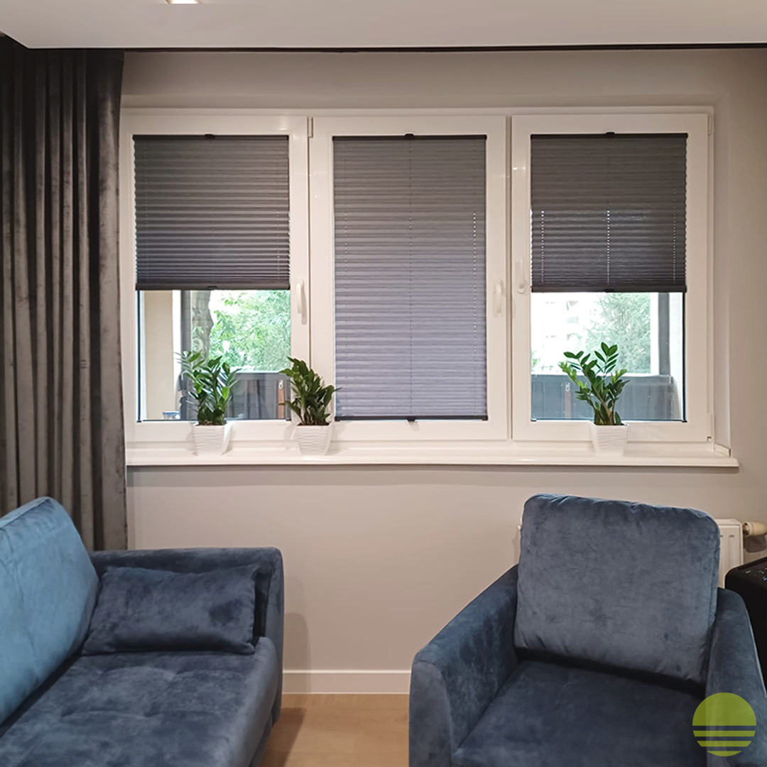 Trzy okna PCV z szarymi plisami, każda z doniczką z zieloną rośliną na parapecie, w tle zasłona i fragment niebieskiej sofy.
