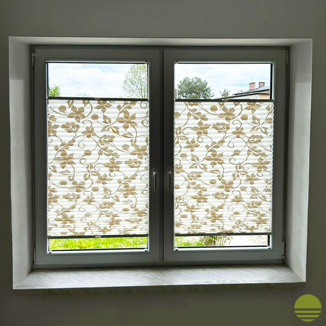 Dwuskrzydłowe okno z białymi ramami, osłonięte plisowanymi roletami z motywem kwiatowym, widok na zielony trawnik i drzewa.
