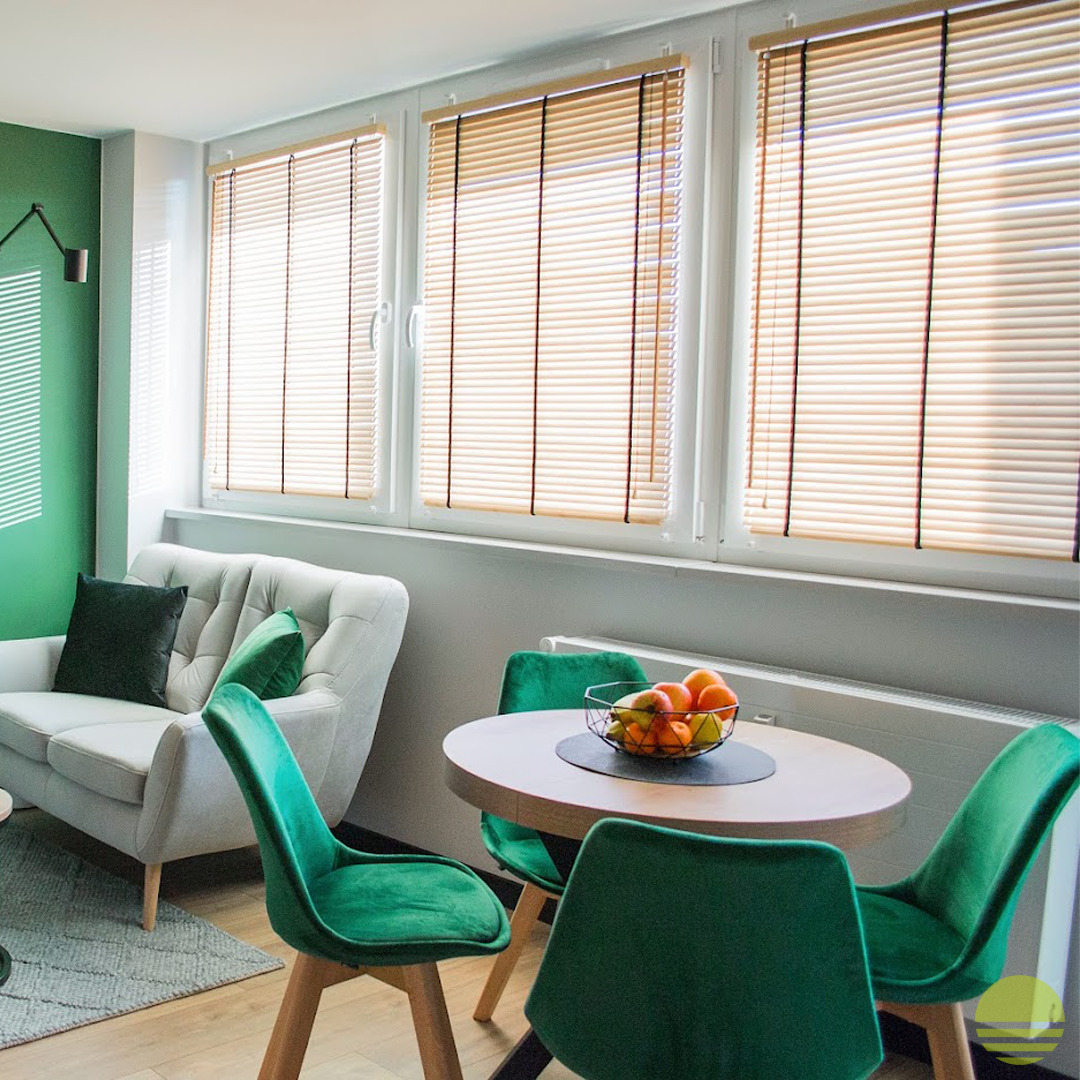 Wnętrze pokoju z trzema oknami z beżowymi żaluzjami poziomymi, szarym narożnikiem, okrągłym stołem i zielonymi krzesłami.