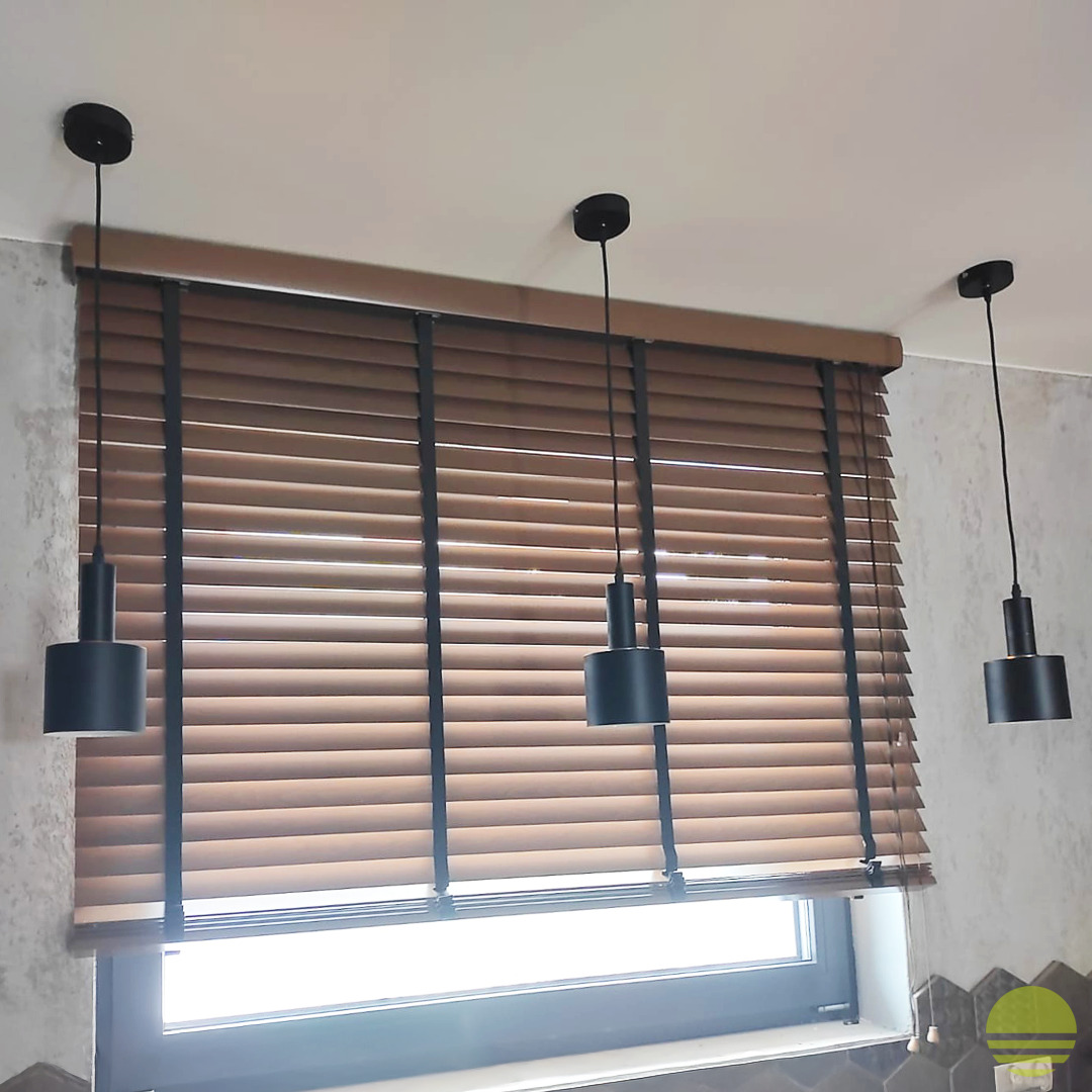 Brązowe poziome osłony okienne z czarnymi taśmami, zamontowane w oknie z trzema czarnymi lampami wiszącymi na górze. Widoczny fragment ściany z szarą teksturą.