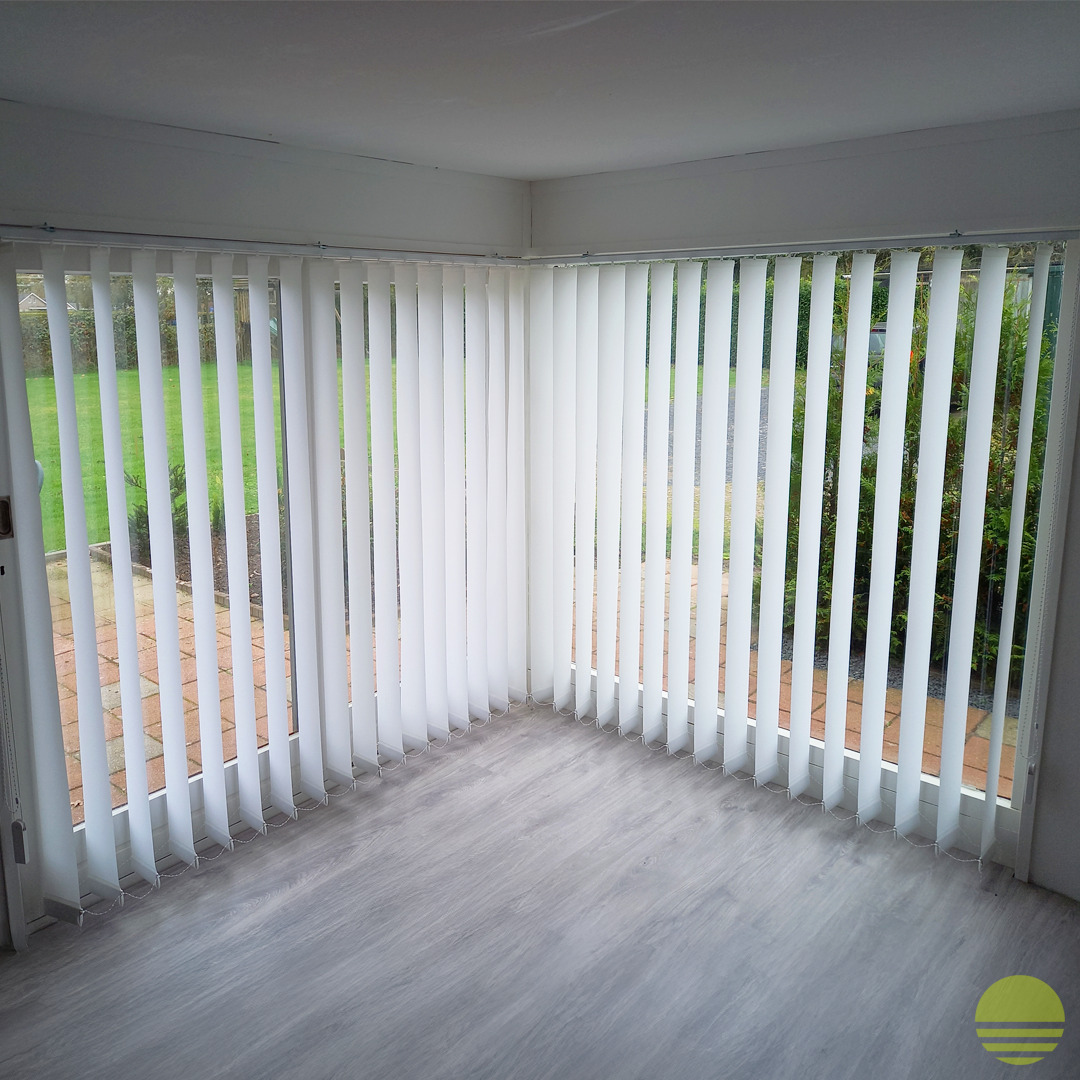 Wnętrze jasnego pomieszczenia z dużymi oknami zasłoniętymi białymi, pionowymi żaluzjami materiałowymi; widok na zielony ogród za oknem.