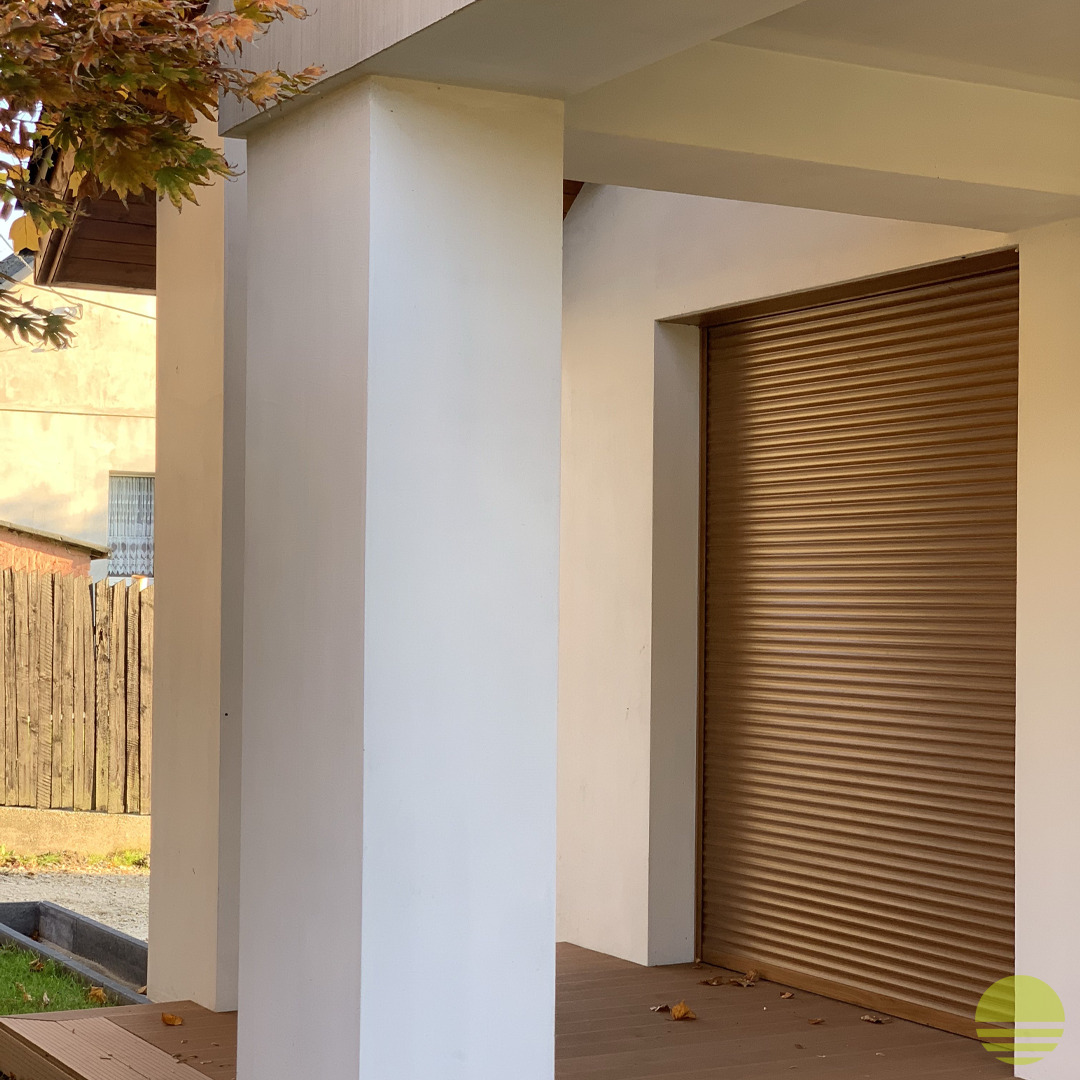 Zewnętrzna roleta antywłamaniowa w kolorze brązowym, zamontowana w nowoczesnym domu z białymi kolumnami, widok z boku w słoneczny dzień.