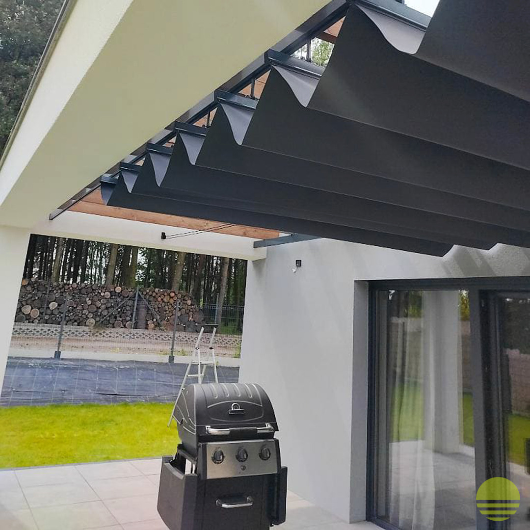 Pergola z automatycznie rozsuwanym, falistym zadaszeniem w kolorze grafitowym, z grillem na tarasie i stosem drewna w tle.