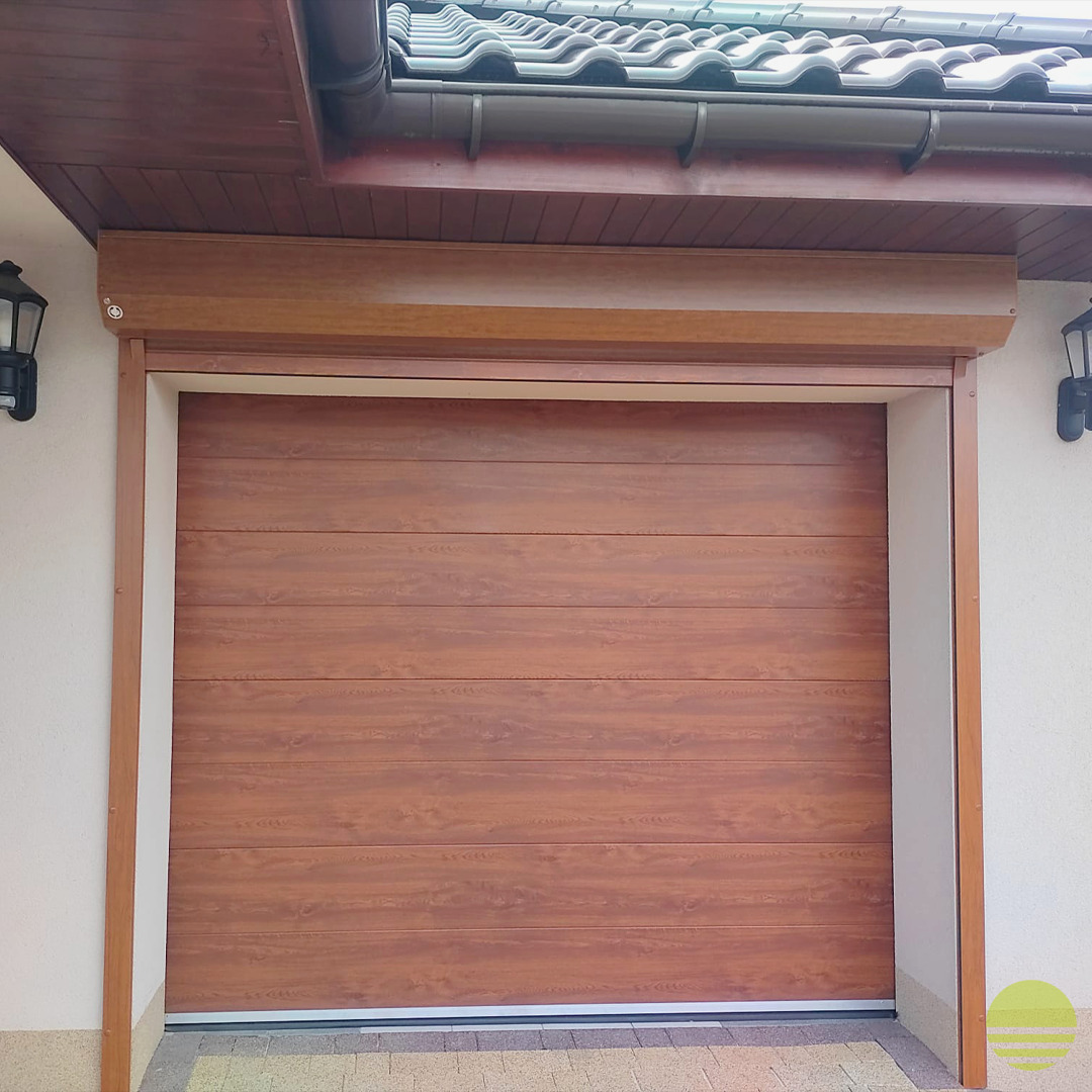 Zewnętrzna brama garażowa segmentowa w kolorze drewna, z widoczną strukturą poziomych paneli, zamontowana w jasnej elewacji budynku z ciemnobrązowym dachem.