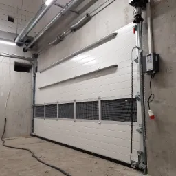 Zamknięta segmentowa brama garażowa z oknami wentylacyjnymi, zamontowana w surowym, betonowym wnętrzu hali przemysłowej, widoczne elementy mechanizmu i sterowania.