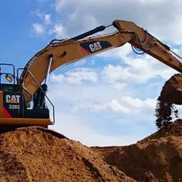 Żółta koparka CAT 320E wysypuje ziemię z łyżki na usypaną górę piasku na tle błękitnego nieba z chmurami.