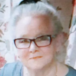 Portret kobiety w okularach z czerwonymi oprawkami, ubranej w bluzkę w kolorze gołębim i naszyjnik z pereł, na tle wzorzystej zasłony.