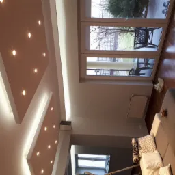 Nowoczesny salon z podwieszanym sufitem z oświetleniem punktowym, widok na taras przez duże okno balkonowe, szara sofa z dekoracyjnymi poduszkami na drewnianej podłodze.