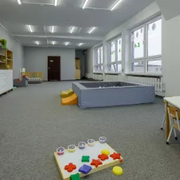 Przestronna sala zabaw z basenem z kulkami, stolikami z krzesłami i regałami z zabawkami, udekorowana naklejkami na ścianach i oknach.