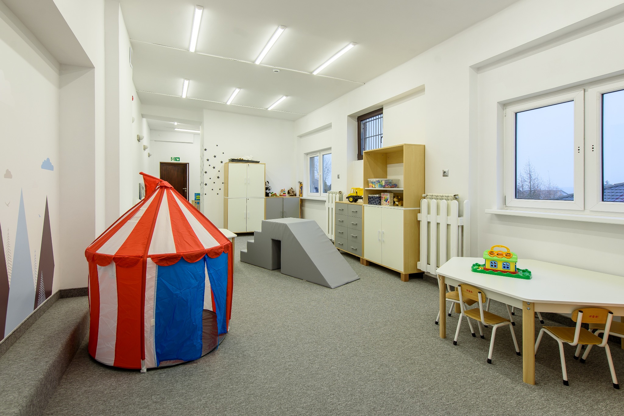 Jasne, przestronne wnętrze sali zabaw z namiotem cyrkowym, miękką konstrukcją do wspinania, stolikiem z krzesełkami i regałami z zabawkami.