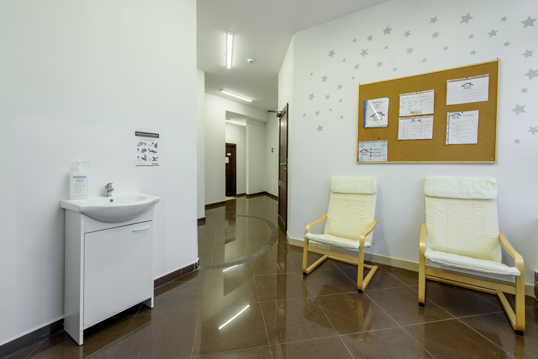 Wnętrze placówki w Katowicach: umywalka z dozownikiem mydła, instrukcja higieny rąk, tablica informacyjna i dwa krzesła w korytarzu z brązowymi płytkami.