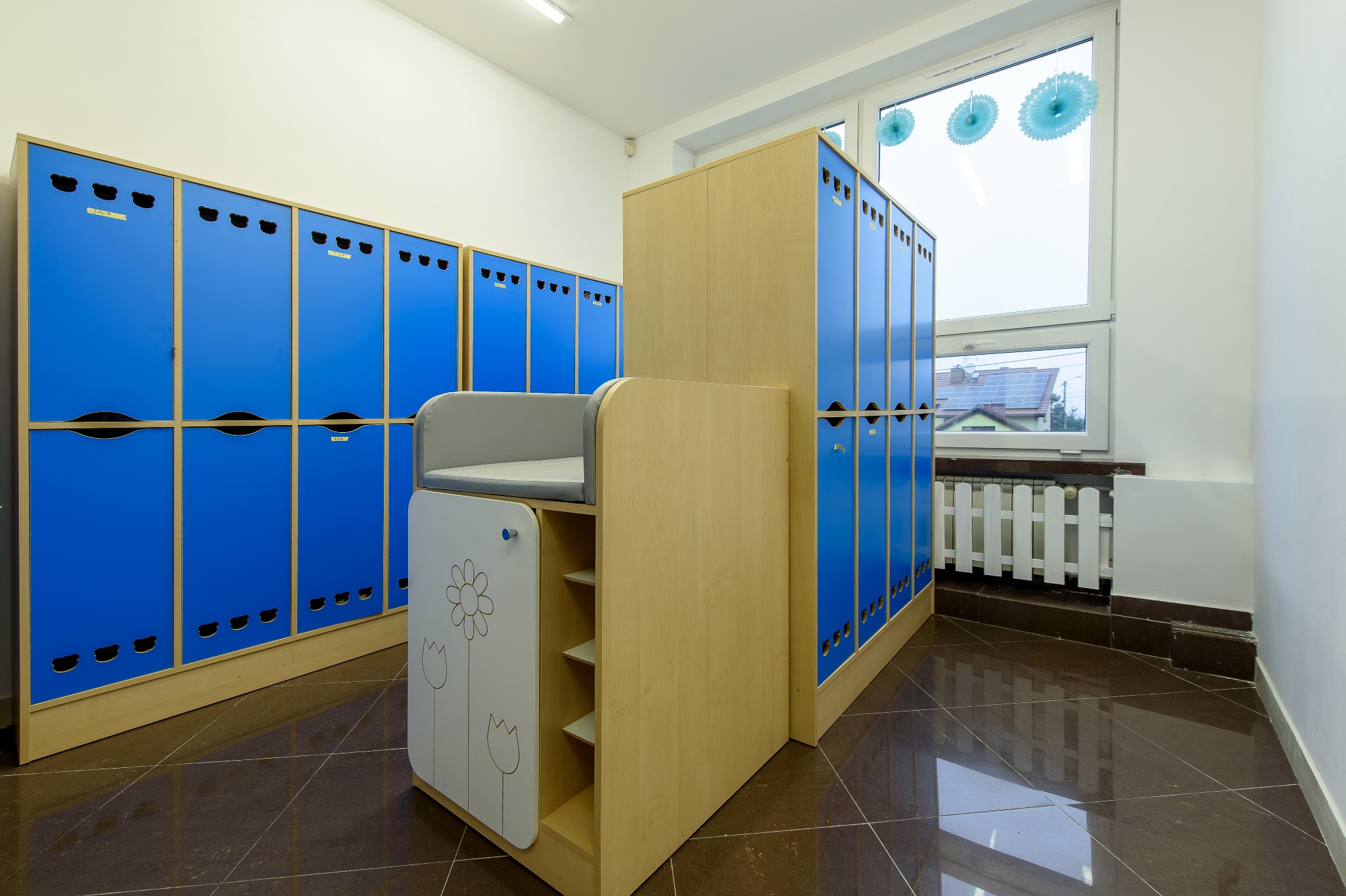 Jasne wnętrze szatni z rzędami niebieskich szafek z wyciętymi motywami zwierząt i niską szafką z przewijakiem ozdobioną rysunkami kwiatów.