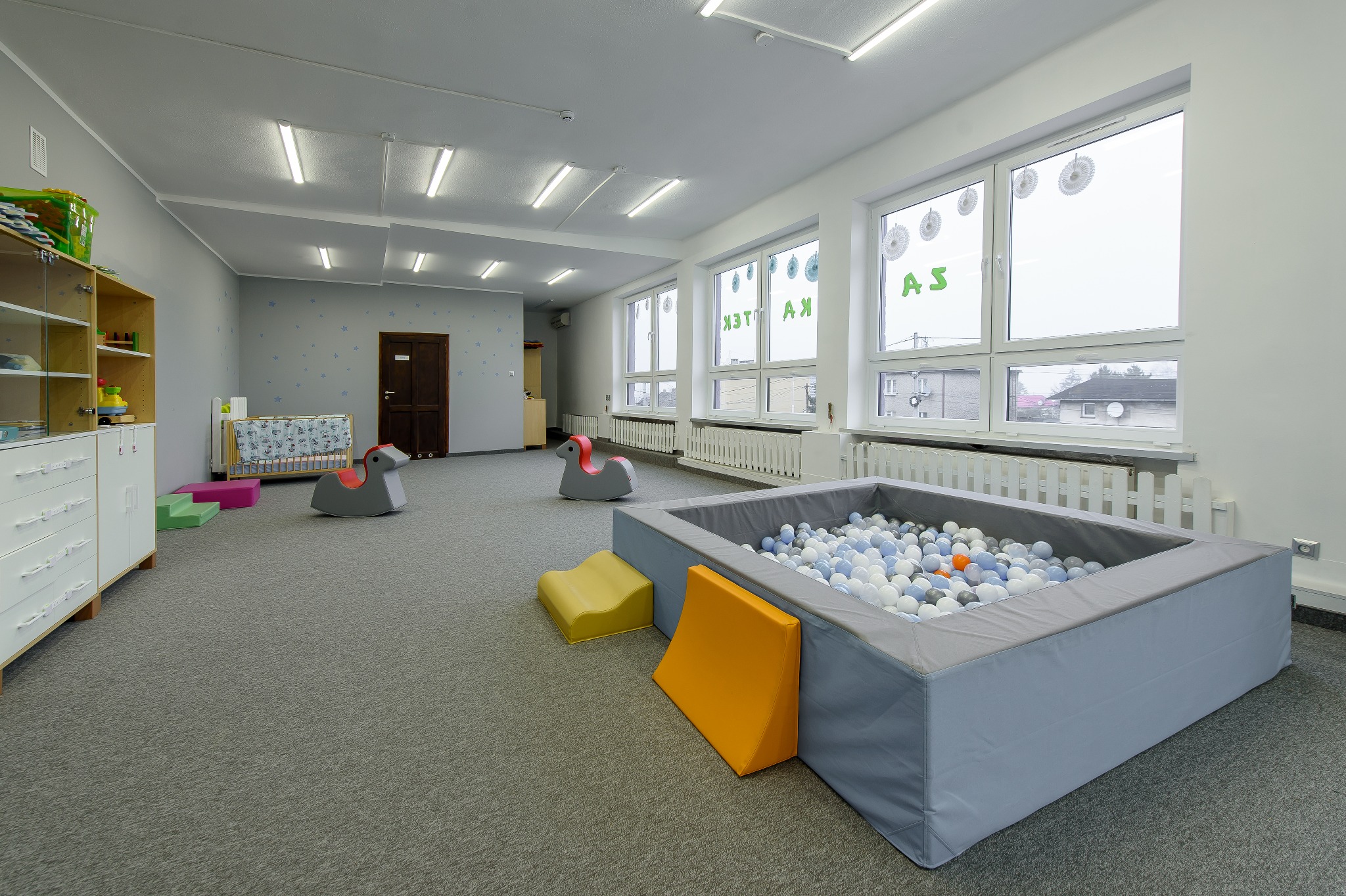 Jasna sala zabaw z basenem z kulkami, drewnianym łóżeczkiem, zabawkami konikami na biegunach i szafkami, udekorowana gwiazdkami na ścianie i naklejkami na oknach.