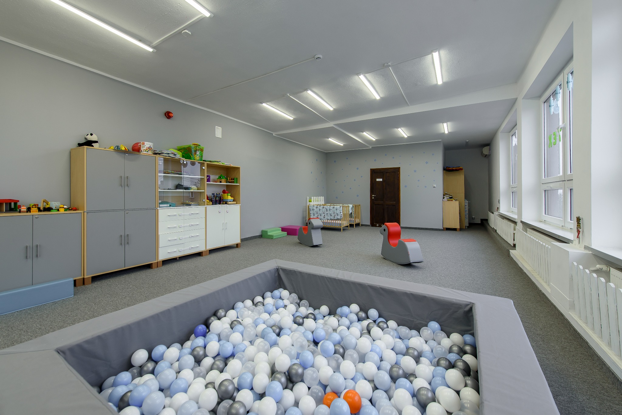 Sala zabaw w żłobku z basenem wypełnionym kulkami w odcieniach bieli, szarości i błękitu, wyposażona w szafki na zabawki, kołyskę i bujaki dla dzieci.