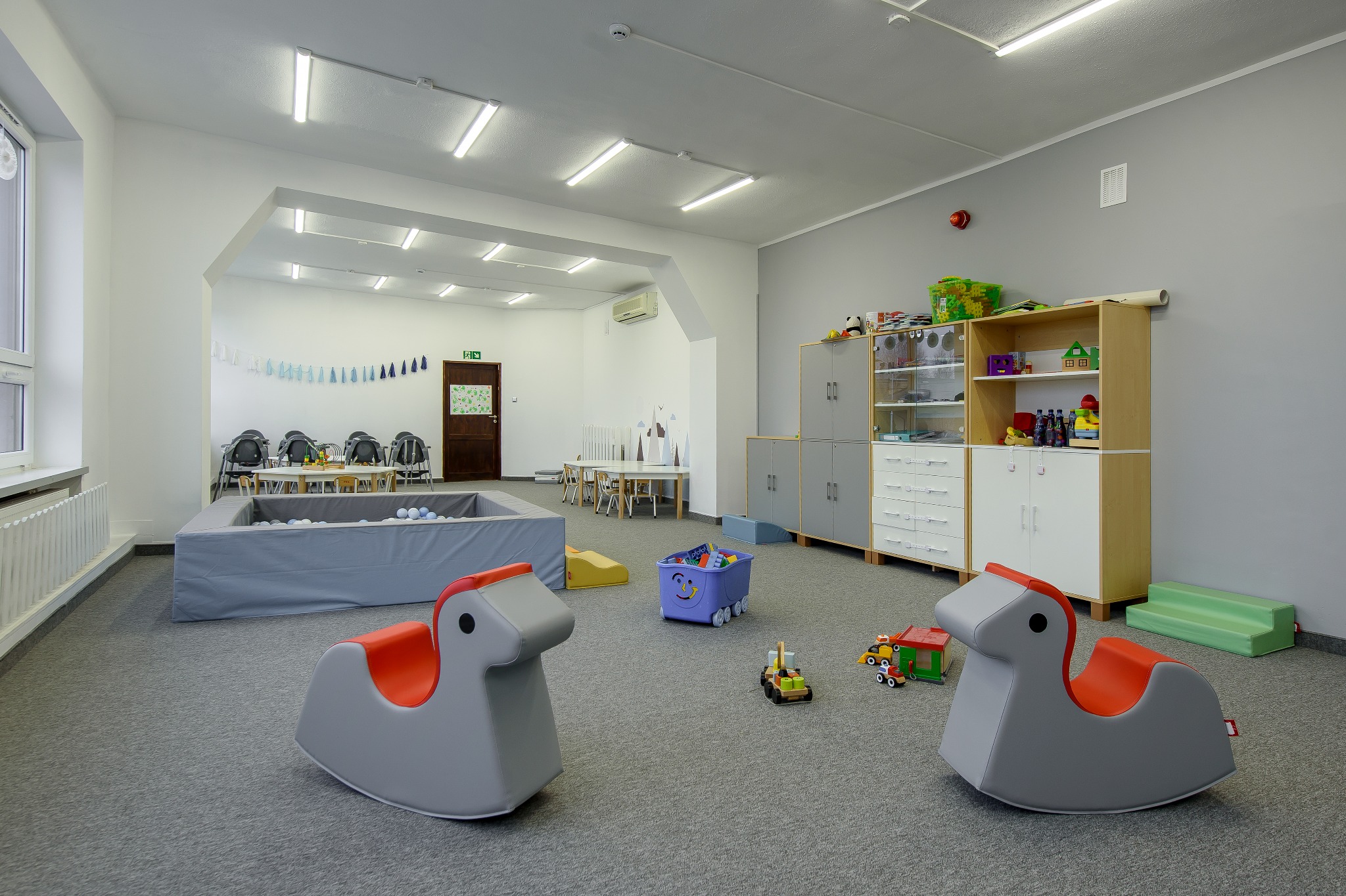 Jasne, przestronne wnętrze sali zabaw z basenem z kulkami, bujakami w kształcie koników i regałami z zabawkami.
