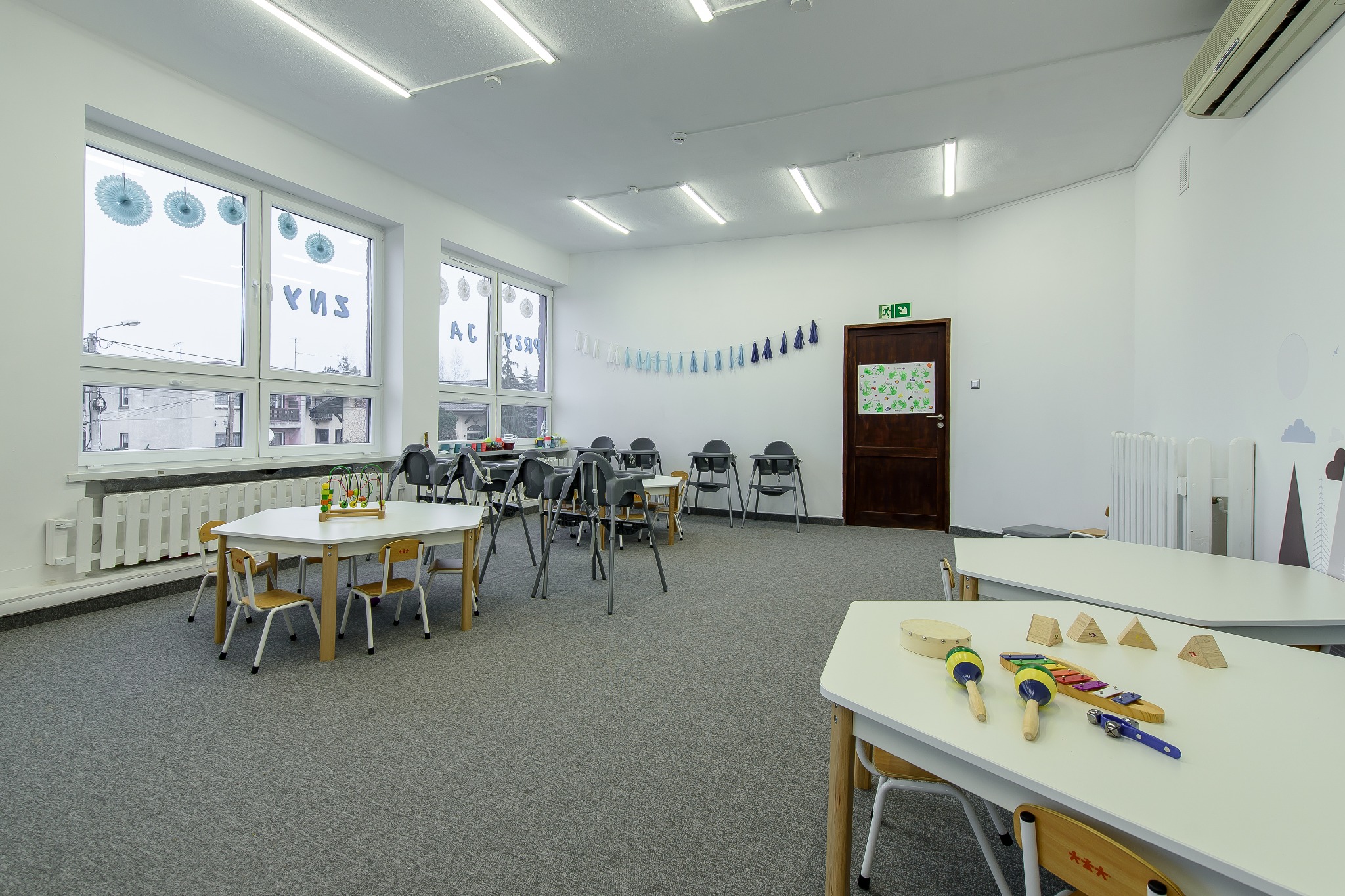 Jasne wnętrze sali zabaw z białymi ścianami, szarym dywanem, stolikami z krzesełkami i zabawkami edukacyjnymi, okna z dekoracjami i napisami, oraz dekoracyjną girlandą pod sufitem.