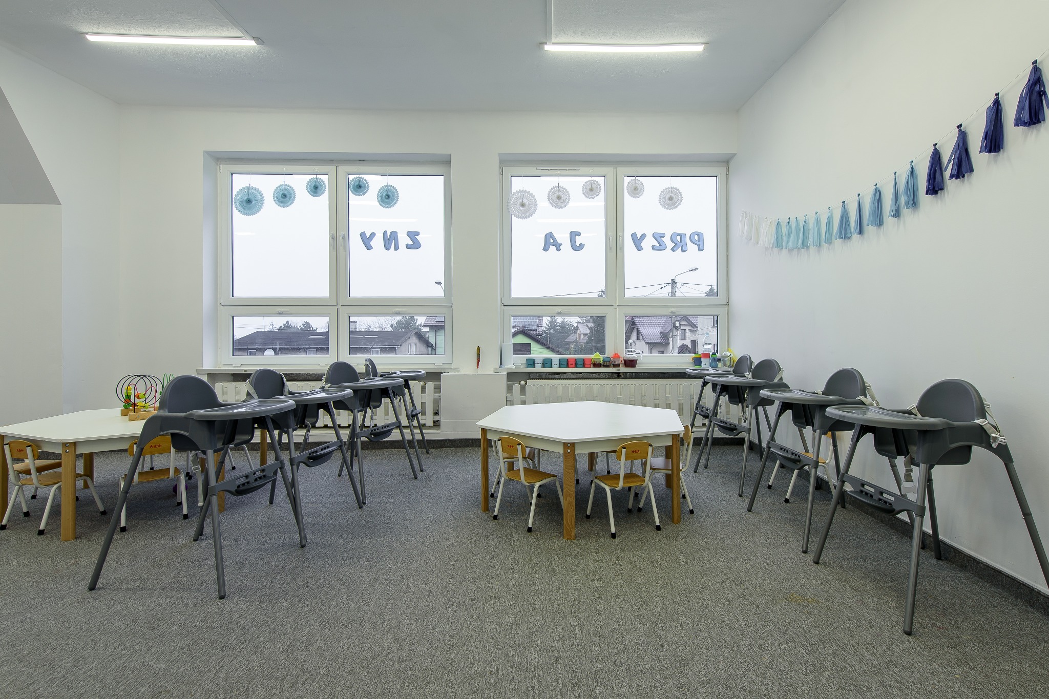 Jasne pomieszczenie z białymi ścianami, oknami ozdobionymi papierowymi dekoracjami i napisami, wyposażone w małe stoliki i szare krzesełka do karmienia, tworzące przestrzeń edukacyjną.