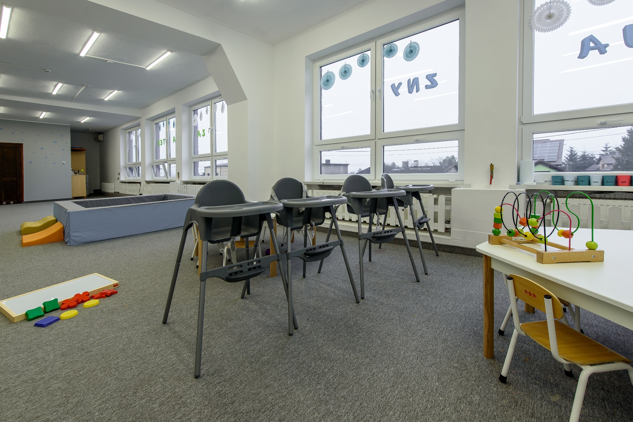 Jasne, przestronne wnętrze sali zabaw z wysokimi krzesełkami, stolikiem z zabawkami edukacyjnymi i basenem z kulkami w tle.