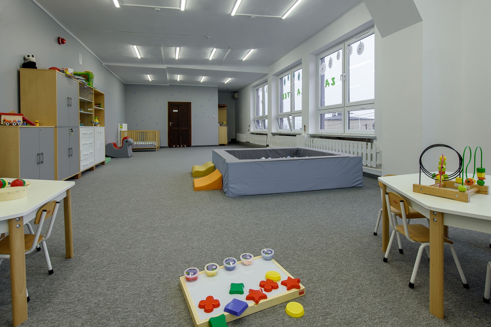 Przestronna sala zabaw z basenem z kulkami, stolikami z krzesłami i regałami z zabawkami, udekorowana naklejkami na ścianach i oknach.