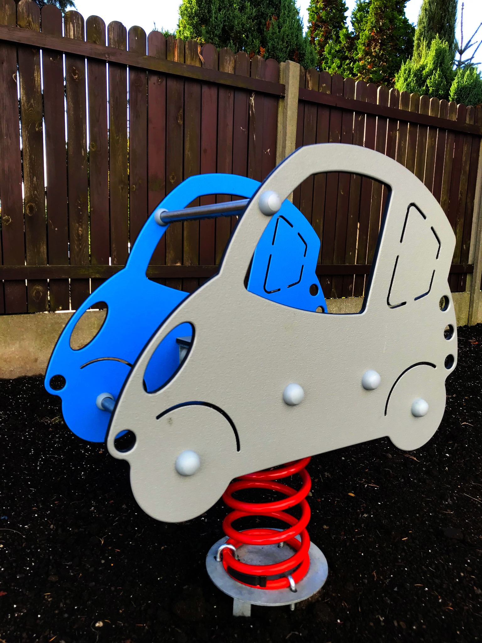 Dwa metalowe samochodziki na sprężynie, jeden niebieski, drugi szary, stojące na czarnej ziemi z drewnianym płotem w tle.
