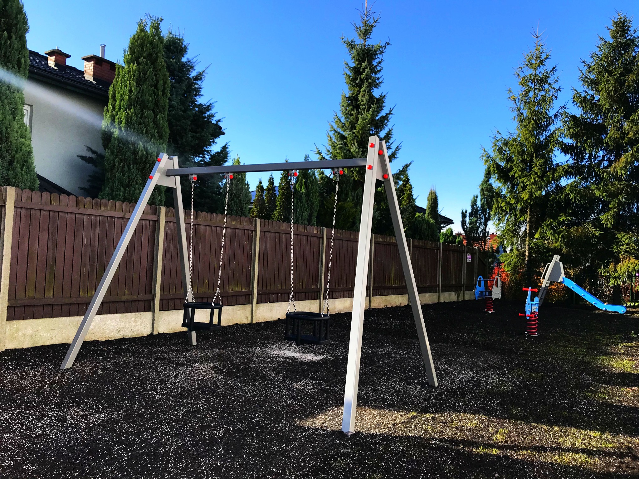 Nowoczesny plac zabaw z huśtawkami kubełkowymi, sprężynowcami i zjeżdżalnią na czarnej, bezpiecznej nawierzchni, otoczony drewnianym płotem i zielenią.