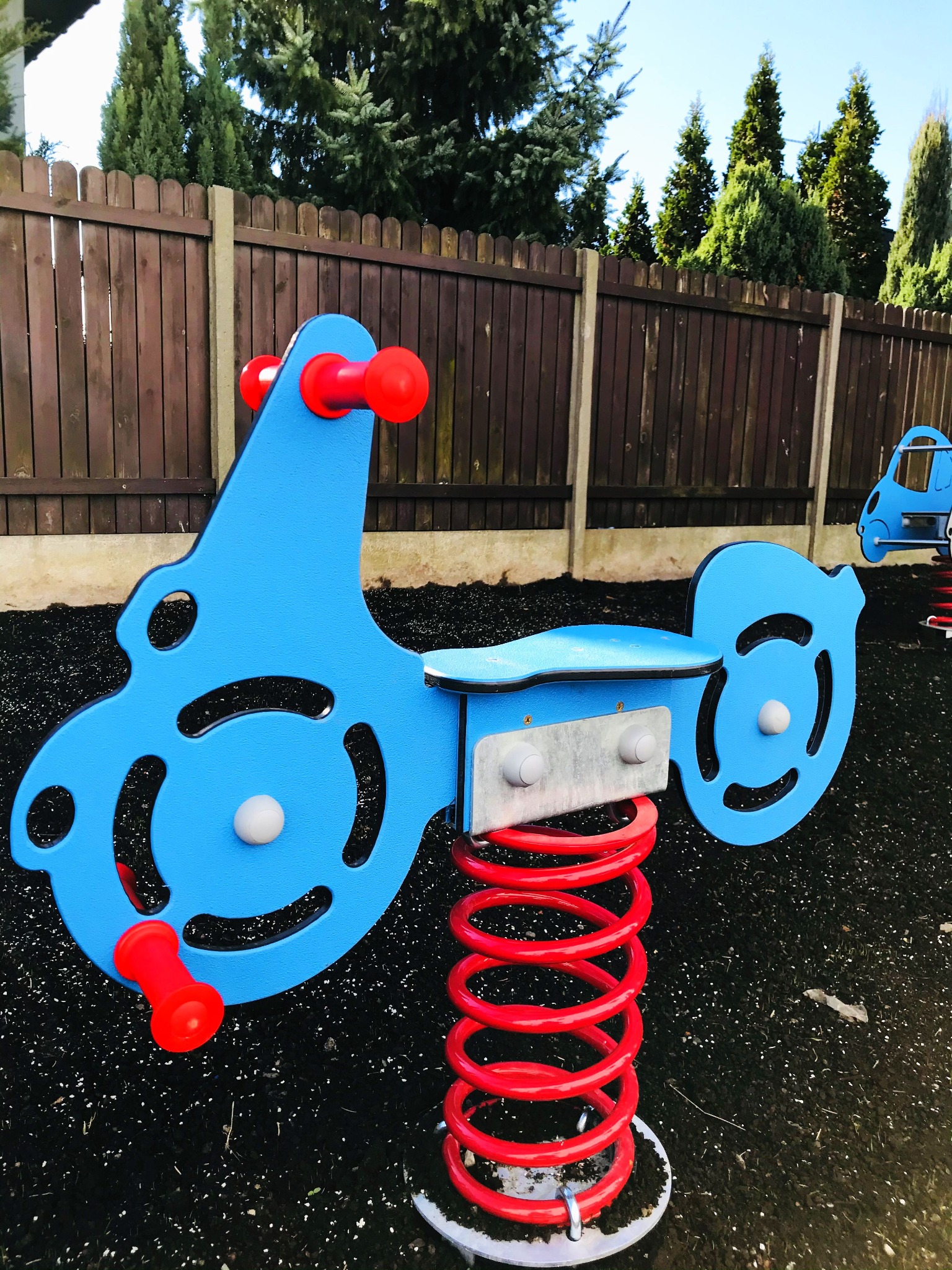 Niebieski bujak sprężynowy w kształcie pojazdu z czerwonymi uchwytami na czarnej nawierzchni placu zabaw, na tle drewnianego płotu i zielonych drzew.