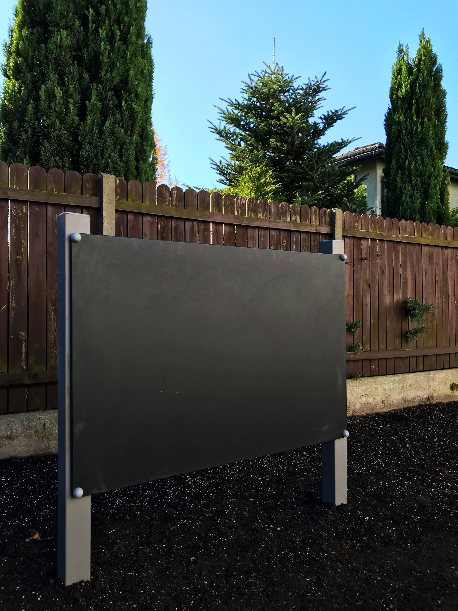 Tablica informacyjna na szarych słupkach na tle brązowego płotu i zielonych drzew iglastych pod błękitnym niebem.