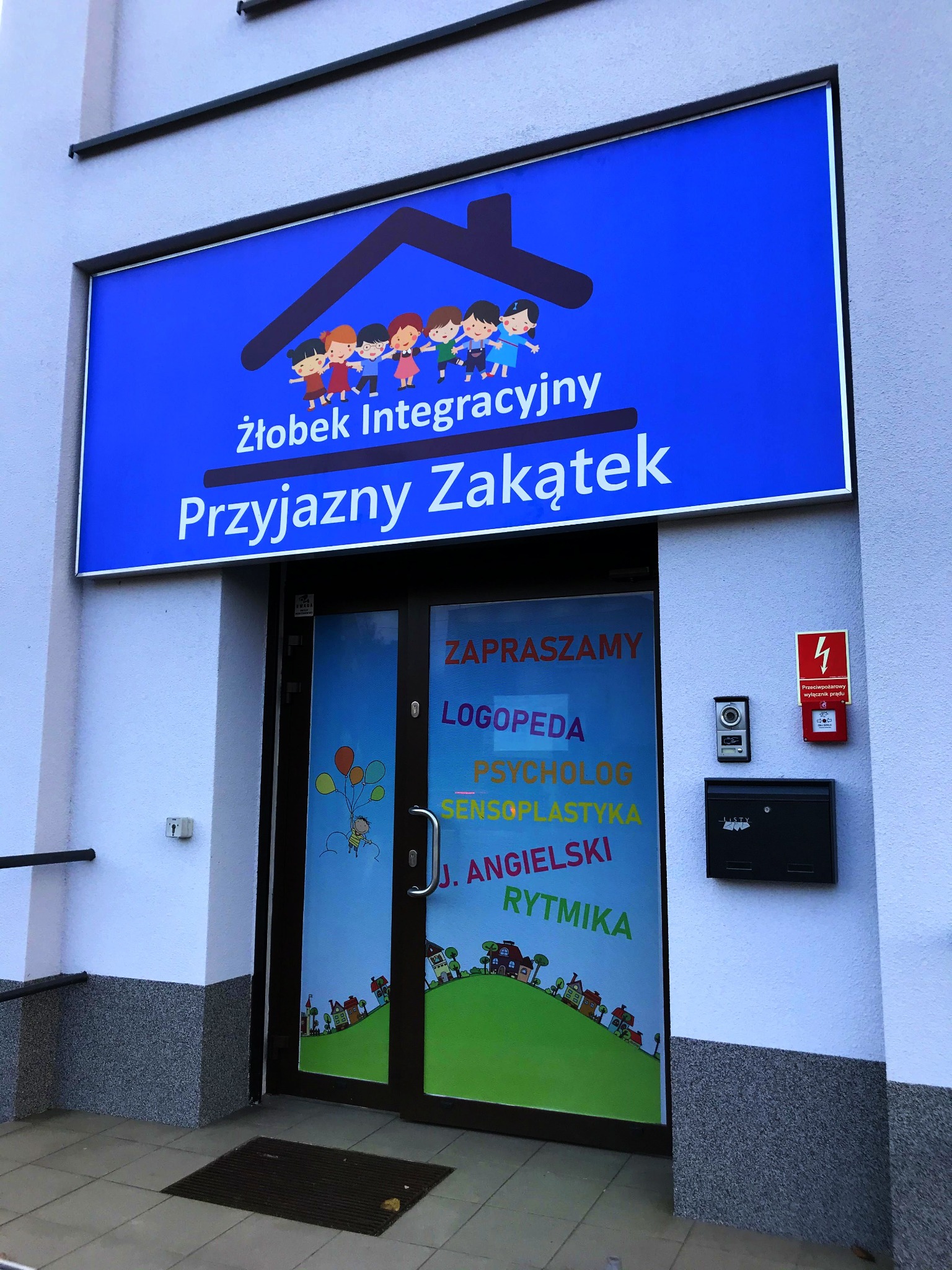 Wejście do placówki z szyldem 'Żłobek Integracyjny Przyjazny Zakątek', na drzwiach naklejki z ofertą: logopeda, psycholog, sensoplastyka, j. angielski, rytmika, na ścianie skrzynka na listy...