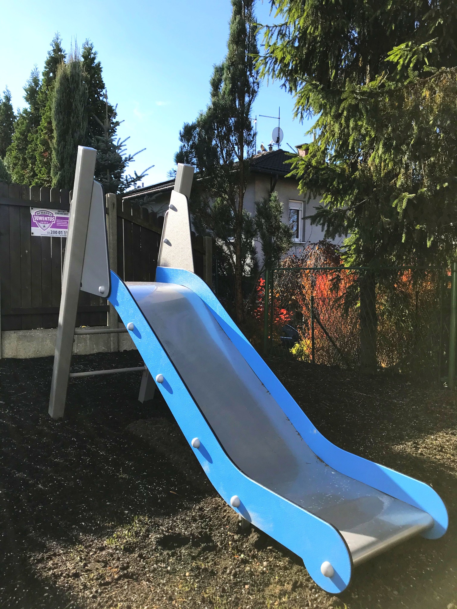 Niebiesko-szara zjeżdżalnia na placu zabaw z widocznym ogrodzeniem i budynkiem mieszkalnym w tle.