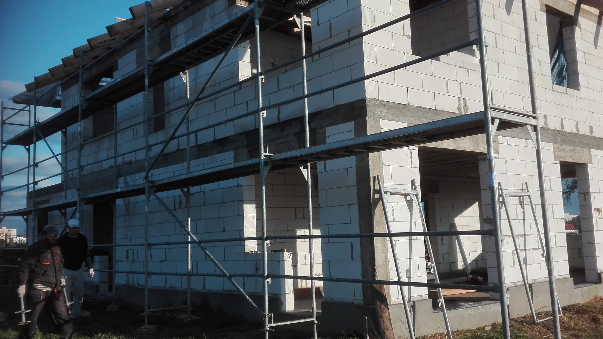 Budowa domu jednorodzinnego z białych bloczków, widoczne rusztowania, dwóch pracowników w odzieży roboczej i narzędzia, stan surowy otwarty.