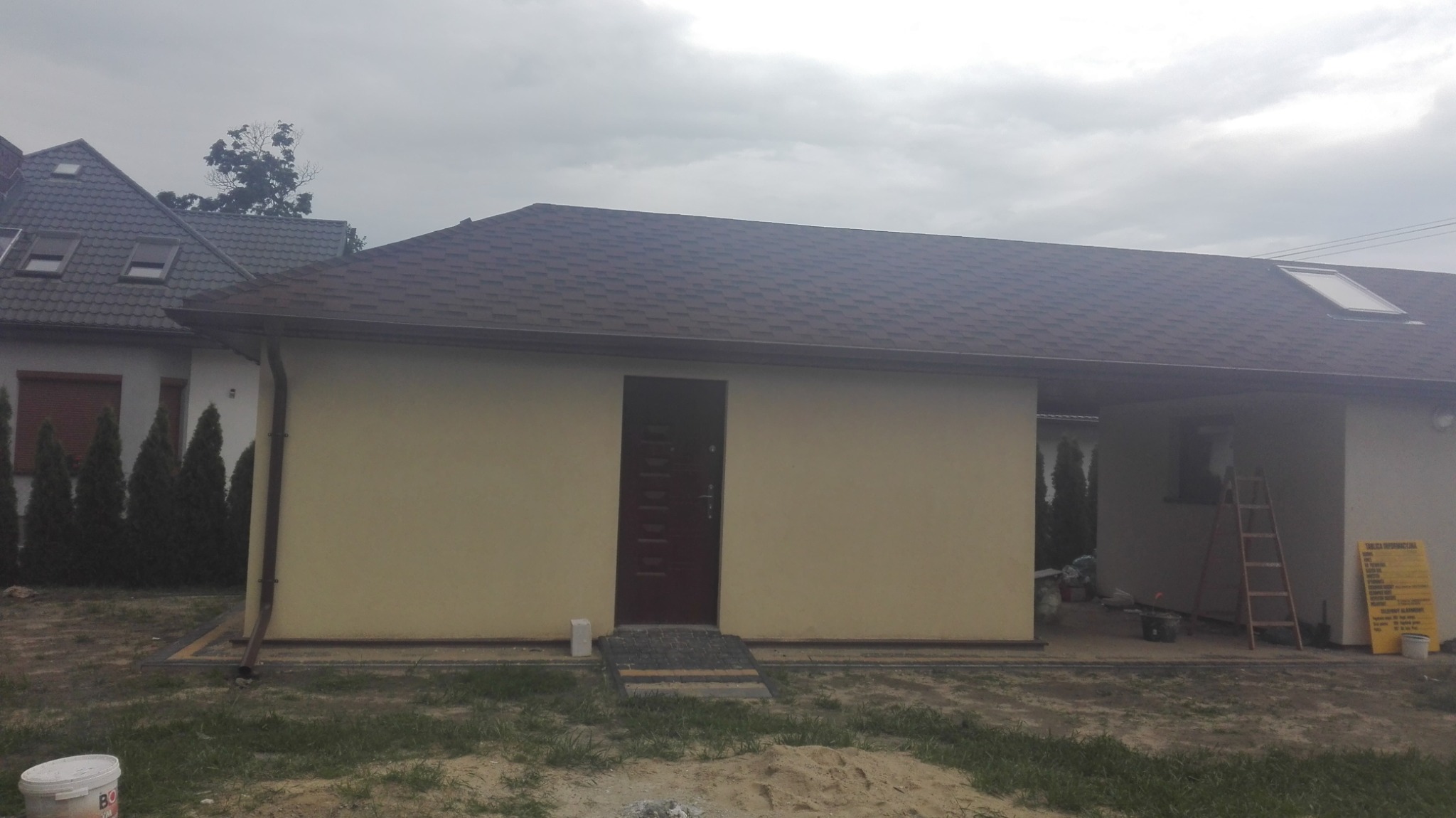 Otynkowany, parterowy dom z brązowym dachem z gontu bitumicznego i ciemnymi drzwiami wejściowymi, widoczny fragment dachu sąsiedniego budynku z oknami dachowymi, pochmurne niebo.