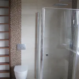 Nowoczesna łazienka z kabiną prysznicową typu walk-in, toaletą podwieszaną i brązowo-beżową mozaiką na ścianie, uzupełniona otwartym regałem z ciemnego drewna.