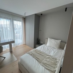 Jasna sypialnia w minimalistycznym stylu: drewniana podłoga, łóżko z białą pościelą i beżowym pledem, biurko przy oknie z białymi zasłonami, szara szafa.