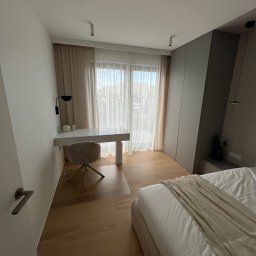 PRO - EXPERT- WYKOŃCZENIA - Jasne wnętrze sypialni z biurkiem, krzesłem i łóżkiem. Parkiet na podłodze, okno z firankami w tle. Minimalistyczny design, stonowana kolorystyka.