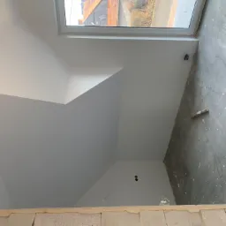Poddasze po świeżym pomalowaniu na biało, widoczne skośne ściany, okno dachowe, surowa wylewka na podłodze z resztkami farby i niedokończony mur poniżej.