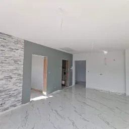 Wnętrze po remoncie: ściana z dekoracyjnej szarej cegły, szara ściana z otworami drzwiowymi, reszta ścian biała, biała marmurowa podłoga, sufit biały z wystającymi kablami.