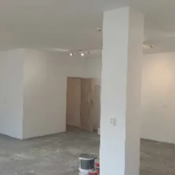 Wnętrze biurowe po świeżym pomalowaniu na biało, widoczne ślady prac na podłodze, puste pomieszczenie z kolumną i lampami sufitowymi.